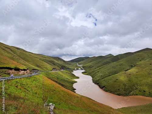 High-angle landscape view of Dishuiyan Viewpoint at Dahai Caoshan (Dahai Grassland) in Huize, Qujing, Yunnan, China. Winding river flowing through lush green rolling hills under cloudy sky.