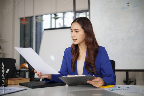Businesswoman reviewing documents and financial reports while sitting at her desk in a modern office.
