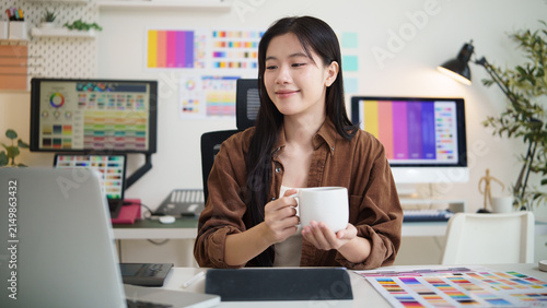 Young female graphic designer holding a coffee mug while working in a modern creative office with colorful design materials.