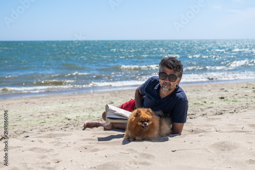 Canvastavla Man and small pomeranian dog lying on sandy beach by the sea, relaxing and readi