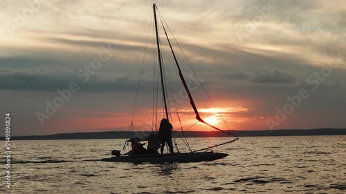 A crewed sailing catamaran moves across a lake at sunset, in slow motion.