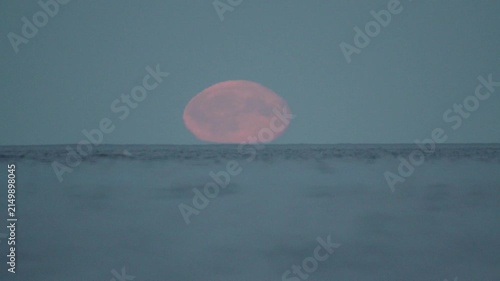 a large red moon is rising above the water surface, in slow motion.