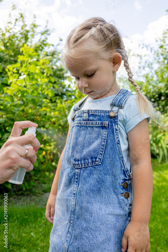 Parent spraying insect repellent on little girl clothes outdoors in garden.