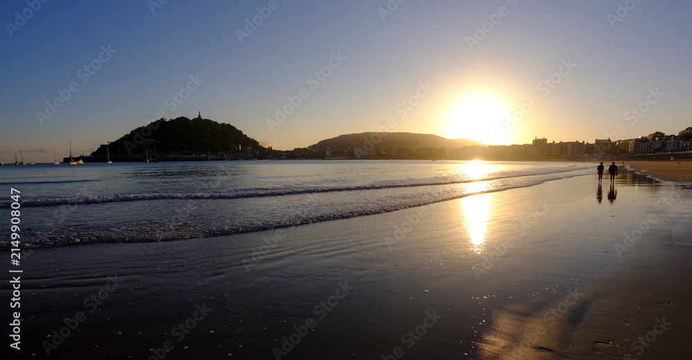 Fototapeta premium Couple walking at sunrise in La Concha Bay, San Sebastian Donostia, Basque Country, Spain. Scenic view of Urgull mountain and city skyline.