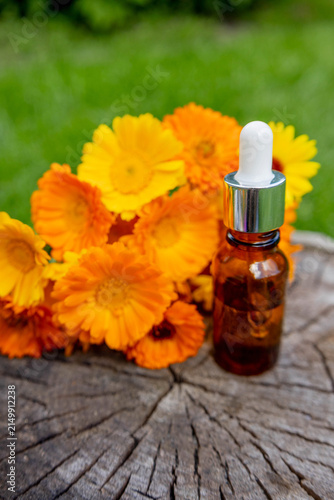 Calendula essential oil bottle and dropper on tree stump with marigold flowers.