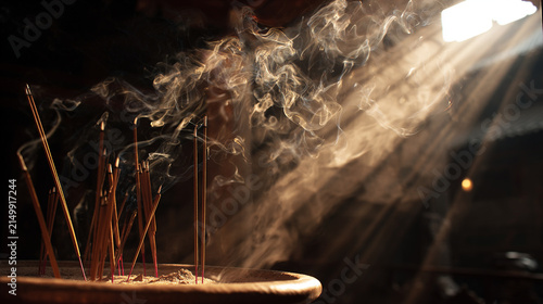 Burning incense sticks in a bronze censer with thick smoke illuminated by sunlight.