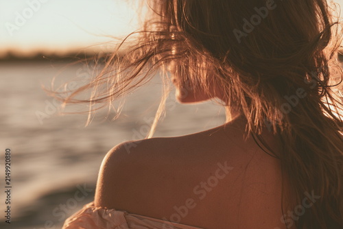 Warm sunset portrait of a woman by the water, with windblown hair and golden light, creating a dreamy summer mood of freedom, calm and quiet escape.