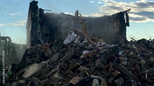 A scene of heavy destruction featuring a collapsed building, piles of rubble, concrete fragments, and dust following a disaster or demolition event.