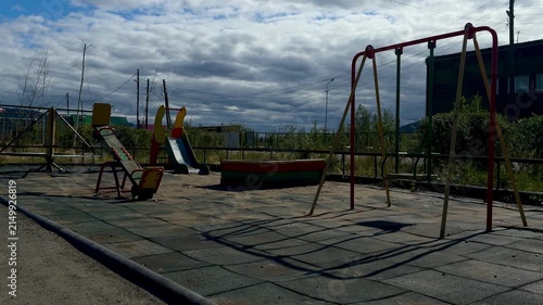 An empty outdoor playground featuring a swing set, a colorful slide, and rubberized safety tiles under a cloudy sky at dusk.
