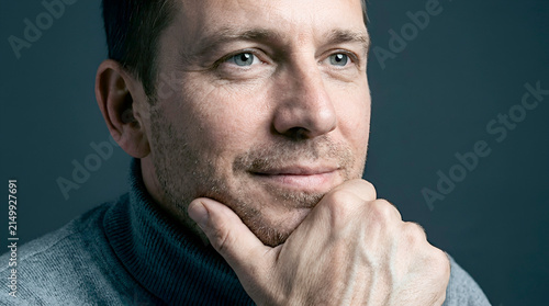 A smiling, attractive middle-aged man in a brown sweater with a genuinely cheerful expression. A close-up studio portrait against a dark background conveys optimism, confidence, and positive emotions.