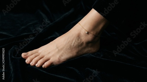 Close up of a barefoot woman's ankle adorned with a delicate gold anklet on a dark velvet background