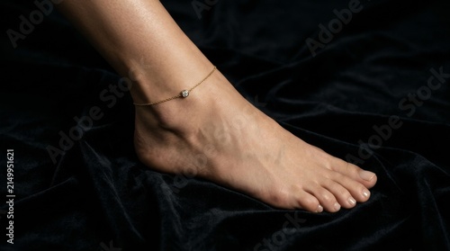 Close up view of a beautiful bare foot adorned with a delicate gold ankle bracelet resting on soft black fabric