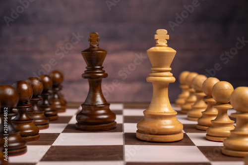 Obraz na płótnie Close-up of wooden chess pieces set up on a chessboard, featuring the white and black kings facing each other with pawns in the background against a dark, textured wall
