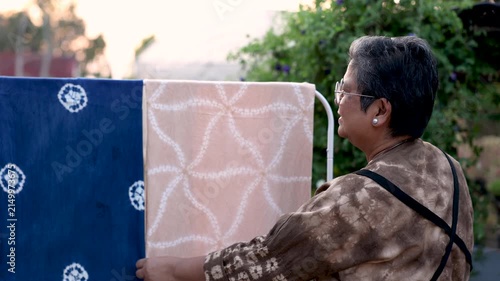 Healthy senior woman arranging colorful dyed fabrics on drying rack outdoors, textile craft business, creative skill, active retirement, productive aging, independent work lifestyle.