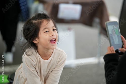 Happy Little Asian Girl Smiling While Being Photographed laughing cheerfully with a smart phone and looking up while someone holds a smartphone in the foreground to take her picture.