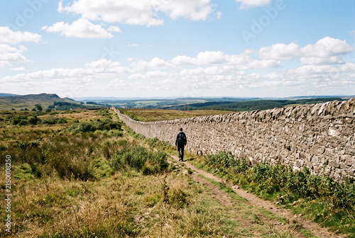 Small distant figure walking alongside long boundary wall in exterior space