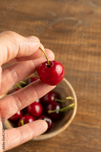 Close-up of hand holding a ripe cherry above wooden bowl filled with cherries on rustic wooden surface, natural fruit photography
