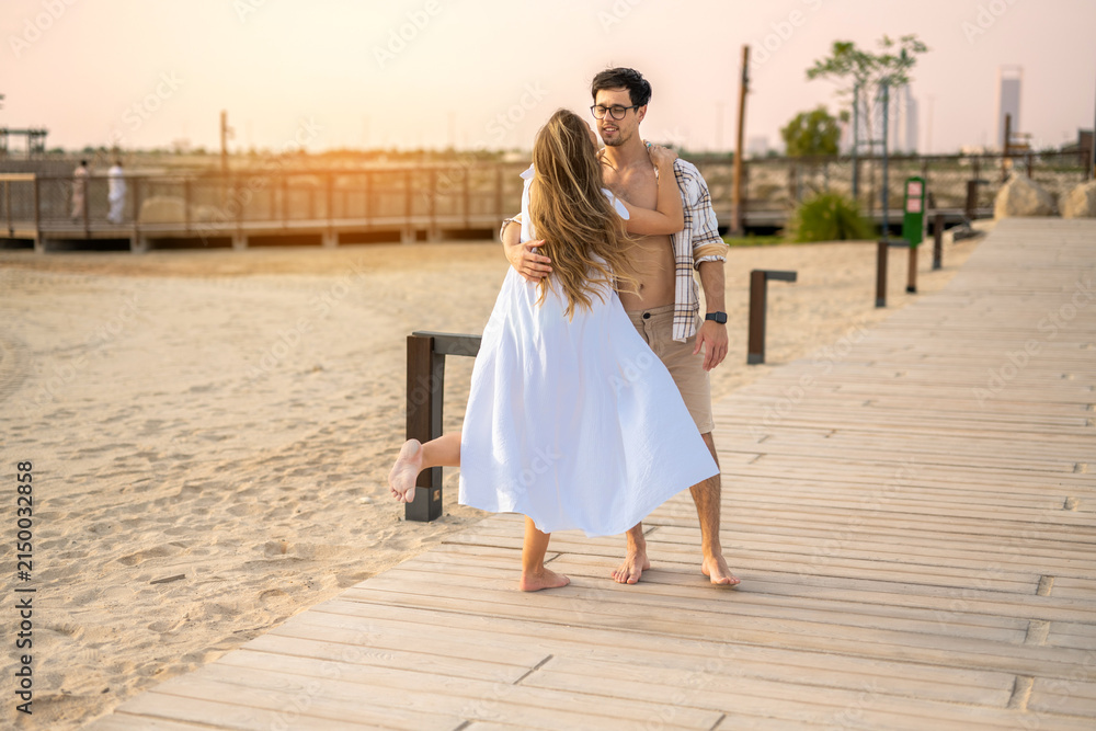 Obraz premium Playful pregnant couple dancing and embracing on a boardwalk by the beach at sunset.