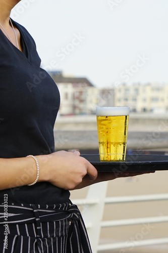 Server holds tray with golden beer. Bubbles rise gently in clear glass. Background hints at waterfront lounge. Neutral expression, focused on service