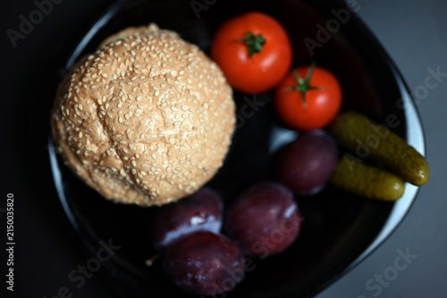 Hamburger with red tomatoes, pickles, and plums on a black background.