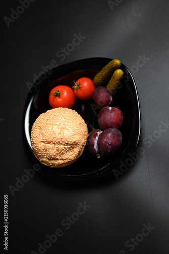 Hamburger with red tomatoes, pickles, and plums on a black background.