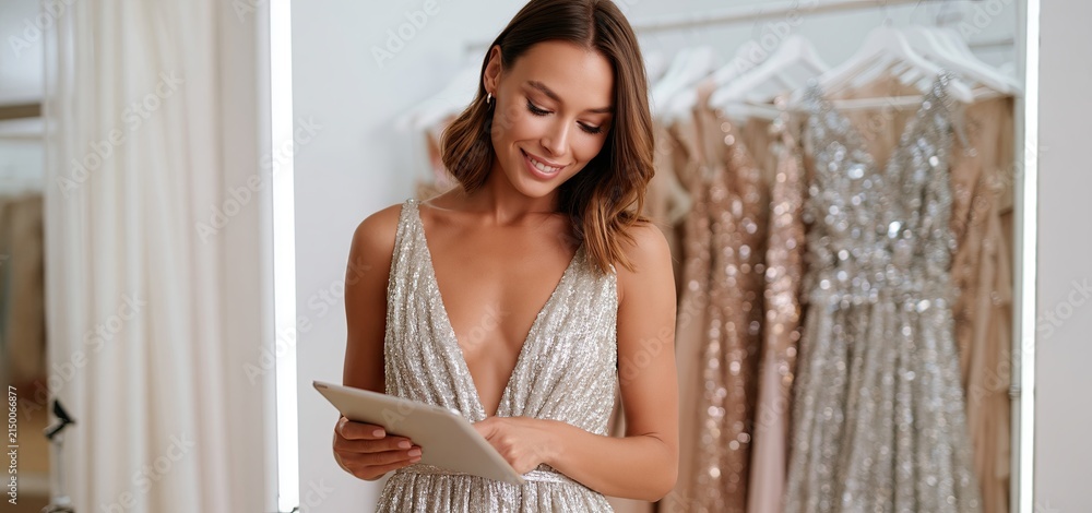Fototapeta premium A fashion consultant selects sparkling gowns from a boutique rack, reviewing options on a tablet in a stylish fitting room.