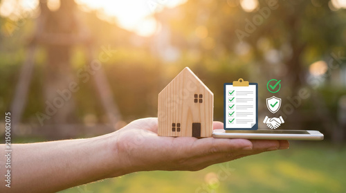 Hand holding house model with quality checklist and approval icon for real estate in natural sunlight
