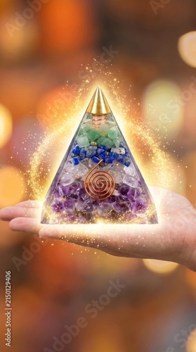 A hand cradling a glowing orgone pyramid adorned with crystals radiating a sparkling aura