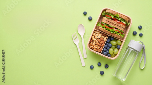 A healthy lunch box containing a fresh sandwich, nuts, grapes, and blueberries, alongside a water bottle and wooden cutlery on a green background.