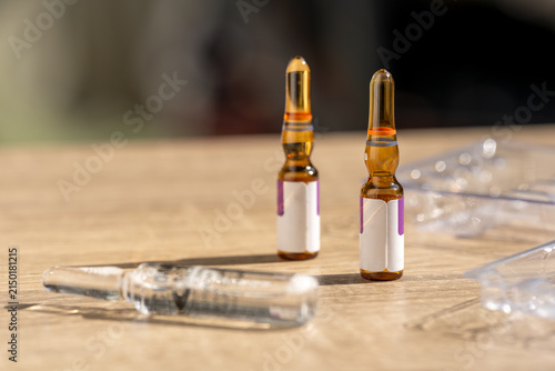 Two amber medication ampoules and a syringe package on a wooden table. Represents injectable treatment, pharmaceutical supplies, clinical medicine and healthcare services in a clean commercial setting
