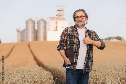 Agronomist holding laptop in wheat field, giving thumbs up, agricultural silos in background, smart farming