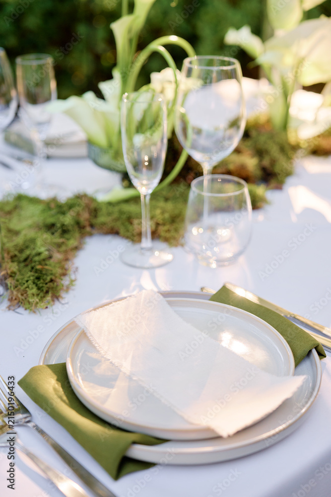 Fototapeta premium Refined outdoor dining scene featuring white tablecloth, stacked plates, crystal glassware, and moss centerpiece. natural light enhance green accents, creating an inviting atmosphere for celebrations