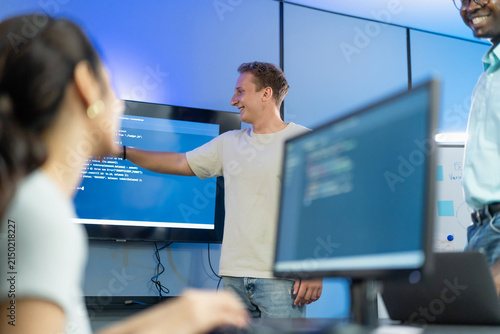 Developer points to and explains code to diverse team members, demonstrating technical knowledge transfer and inclusive group learning through collaborative code walkthrough.