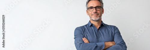 Professional mature businessman in blue button-down shirt standing confidently with arms crossed against white background, displaying friendly smile and modern glasses.