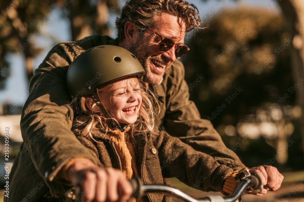 Fototapeta premium Father and daughter laughing together while riding a bicycle outdoors during golden hour