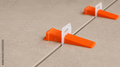 Close-up of tile leveling system with orange wedges and clips installed on grey porcelain floor tiles. Precise alignment process during professional home renovation and construction.