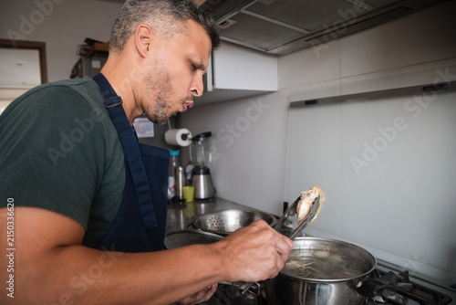 Man Tasting Homemade Broth with Tongs in Kitchen