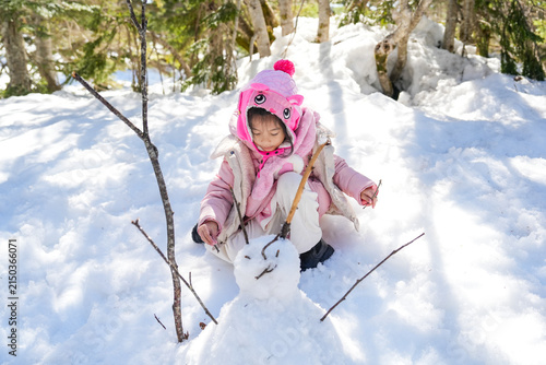 Little Asian Girl pink winter clothes using tree twigs to decorate a miniature snowman in Winter Forest in a forest landscape.