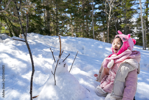 Sun shining to Little Asian Girl face , excited and amazed to snow man open her mouth,she sit down wearing a pink winter jacket winter clothes in snowball in pine Forest.