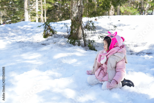 Little Asian Girl look around snow seeing around she sit down wearing a pink winter jacket winter clothes in snowball in pine Forest.