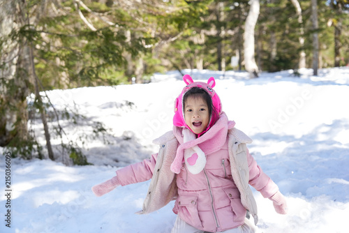 Happy Little Asian Girl wearing a pink winter jacket winter clothes kneeling and Throwing a Snowball in pine Forest.