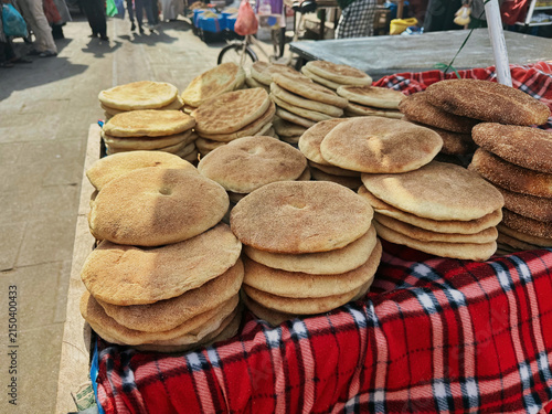 Stacks of freshly baked round Moroccan Khobz Flatbread are displayed for sale on a street market stall covered with a red plaid cloth.