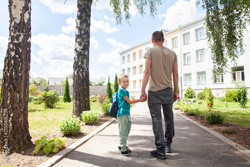 Caring father walks hand in hand with his young son toward the school building on a sunny back to school morning. Child looks back and smiles