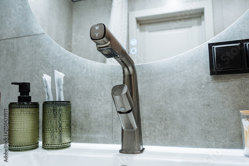 Close-up of modern brushed metal faucet featuring pullout spray head in front of round mirror mounted on gray textured tile wall, with glass soap dispensers, toothbrushes, and clean contemporary