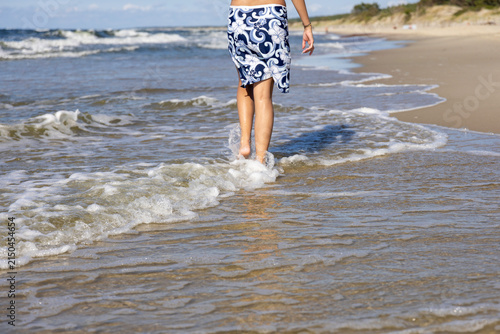A woman in a slit skirt walks barefoot through shallow water, creating splashes, with sandy beach in the background.