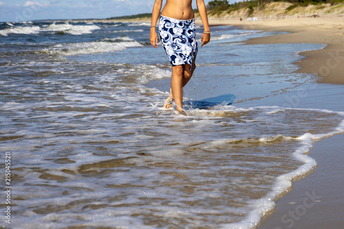 A woman in a slit skirt walks barefoot through shallow water, creating splashes, with sandy beach in the background.