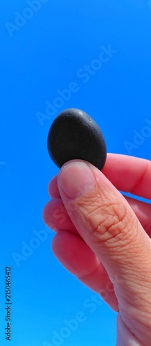 Cuadro en lienzo Hand holding a smooth black oval pebble against a clear vivid blue sky backdrop
