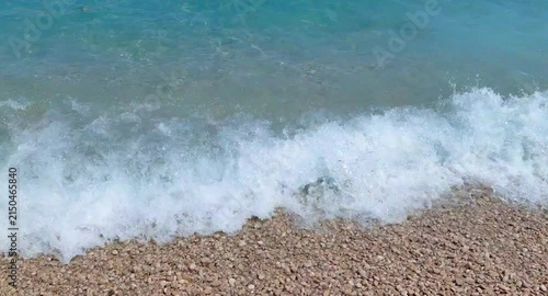 A wide shot of crystal-clear turquoise sea waves rolling onto a pebble beach with white foam under bright summer sunlight, creating a calm and peaceful coastal scene.