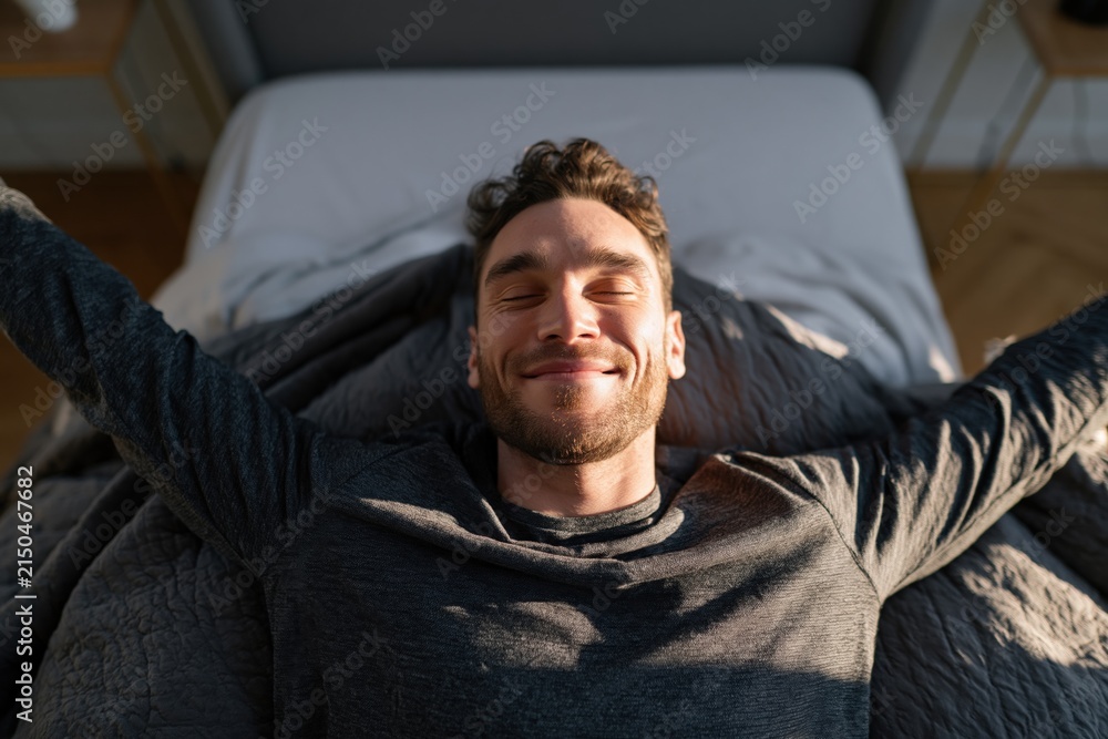 Fototapeta premium Man smiling with sunlight on his face while stretching in bed for morning background wallpaper and wellness banner design