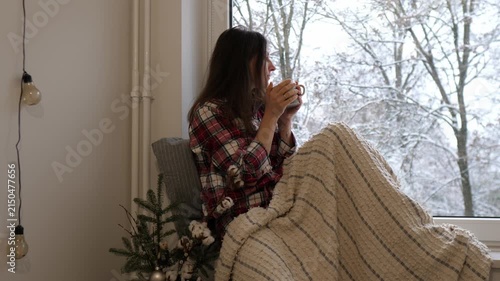 A young woman sitting by the window, wrapped in a warm blanket and holding a cup of hot tea. Snow is falling outside